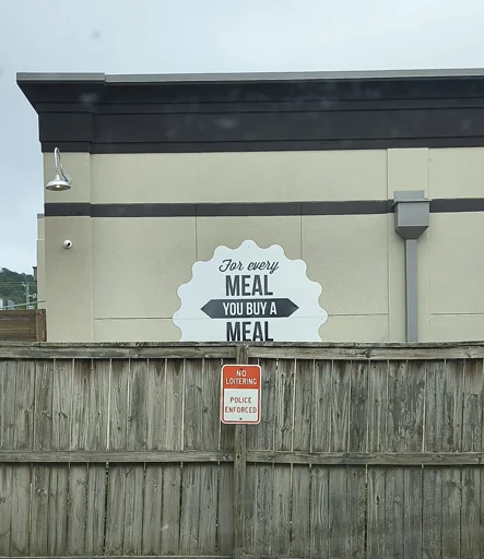 Marketing sticker from a restaurant that donates a meal to impoverished persons for every meal purchased in-store. Half of the sign is cut off by a fence, and what remains reads "FOR EVERY MEAL, YOU BUY A MEAL"